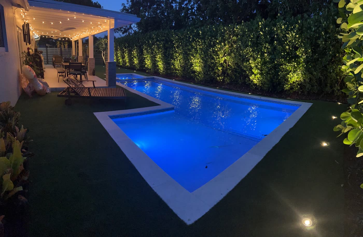 A pool, hedge and covered terrace lit at night